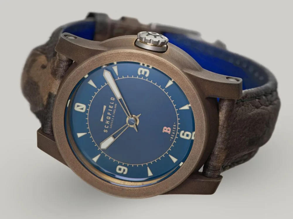 Bronze watches by Schofield Watch Company. Patinated Best Bronze. The B3 ✦ Patinated Bronze wristwatch with a blue dial and brown leather strap. The watch face features bold luminescent numerals at 3, 6, 9, and 12, with smaller indicators for other hours and minutes. The brand name "Schofield" is visible on the dial, along with the Beam of light logo.