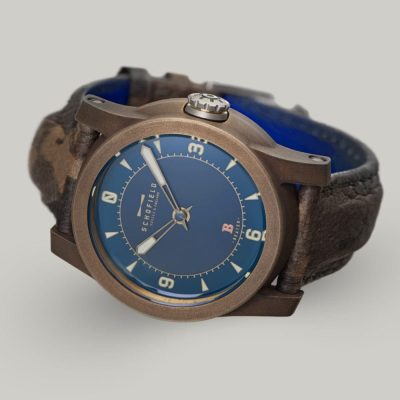 The B3 ✦ Patinated Bronze wristwatch with a blue dial and brown leather strap. The watch face features bold luminescent numerals at 3, 6, 9, and 12, with smaller indicators for other hours and minutes. The brand name "Schofield" is visible on the dial, along with the Beam of light logo.
