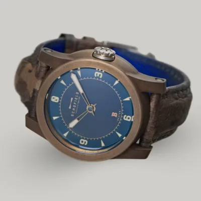 Bronze watches by Schofield Watch Company. Patinated Best Bronze. The B3 ✦ Patinated Bronze wristwatch with a blue dial and brown leather strap. The watch face features bold luminescent numerals at 3, 6, 9, and 12, with smaller indicators for other hours and minutes. The brand name "Schofield" is visible on the dial, along with the Beam of light logo.