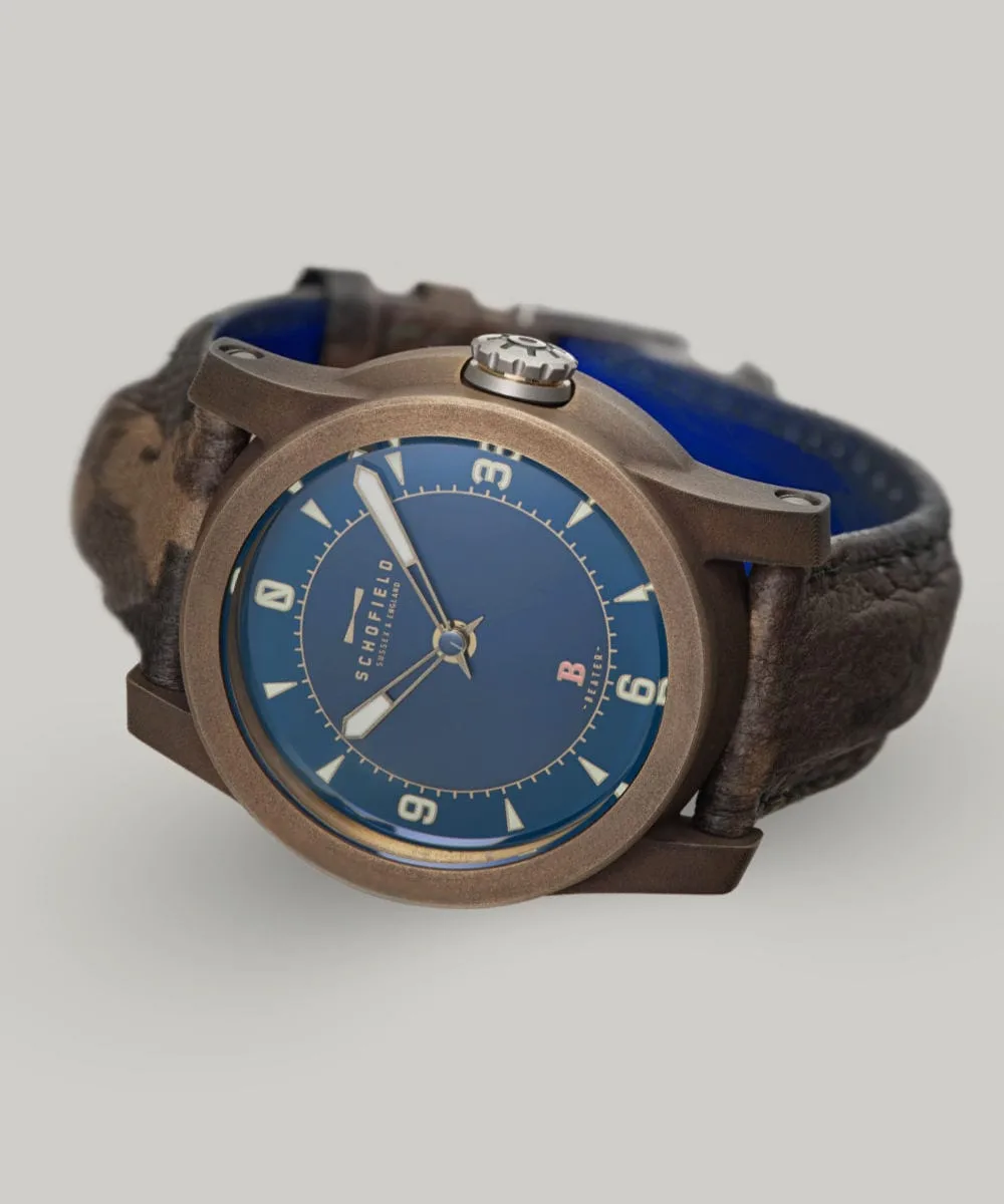 The B3 ✦ Patinated Bronze wristwatch with a blue dial and brown leather strap. The watch face features bold luminescent numerals at 3, 6, 9, and 12, with smaller indicators for other hours and minutes. The brand name "Schofield" is visible on the dial, along with the Beam of light logo.