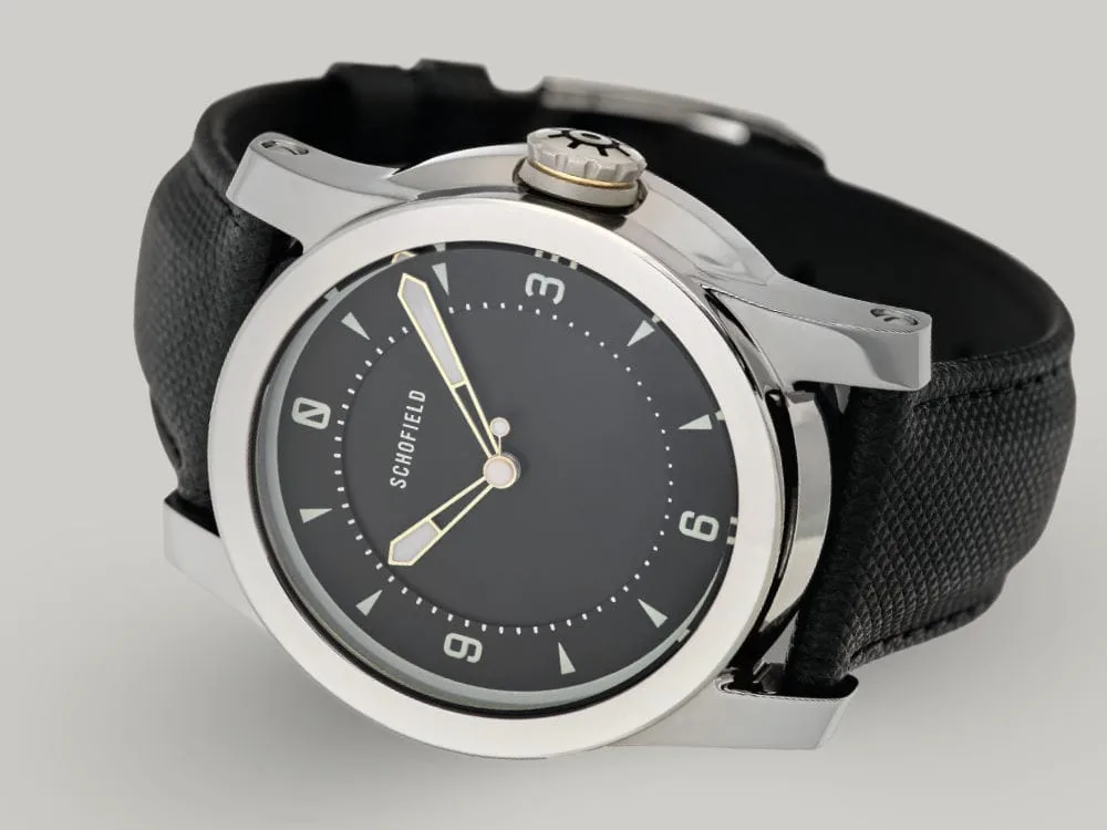 A Treasure Watch ✦ Silver with a stainless steel case and a black leather strap. It features a black dial with white hour markers and hands, along with a crown engraved with a wheel design. Schofield is displayed on the minimal dial.