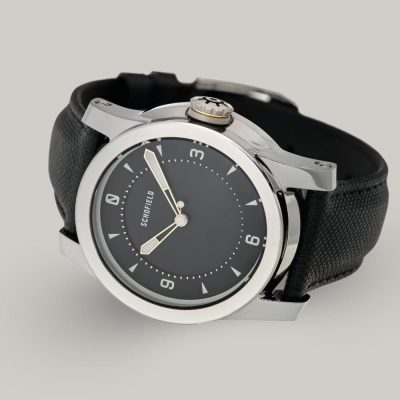 A Treasure Watch ✦ Silver with a stainless steel case and a black leather strap. It features a black dial with white hour markers and hands, along with a crown engraved with a wheel design. Schofield is displayed on the minimal dial.