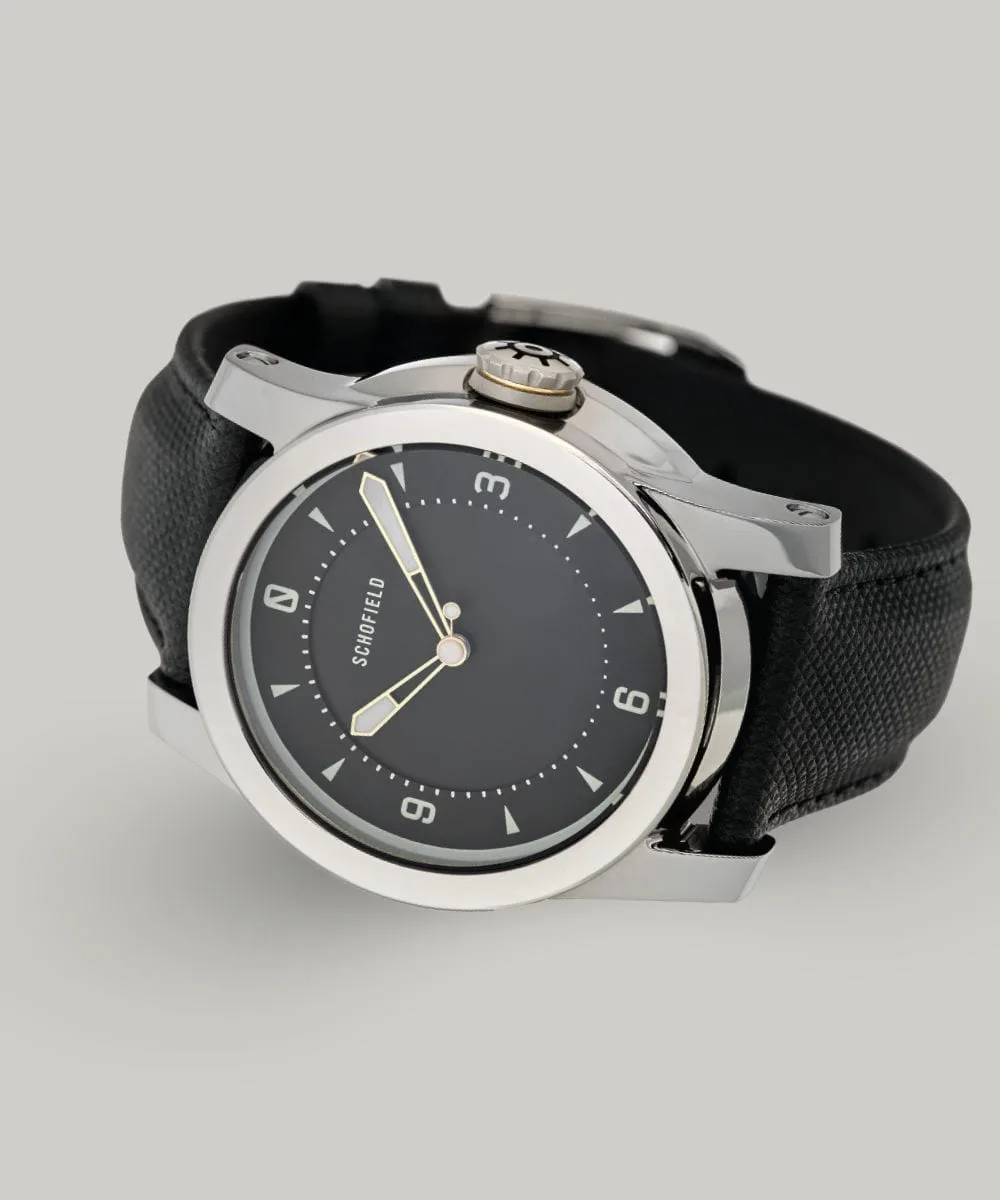 A Treasure Watch ✦ Silver with a stainless steel case and a black leather strap. It features a black dial with white hour markers and hands, along with a crown engraved with a wheel design. Schofield is displayed on the minimal dial.