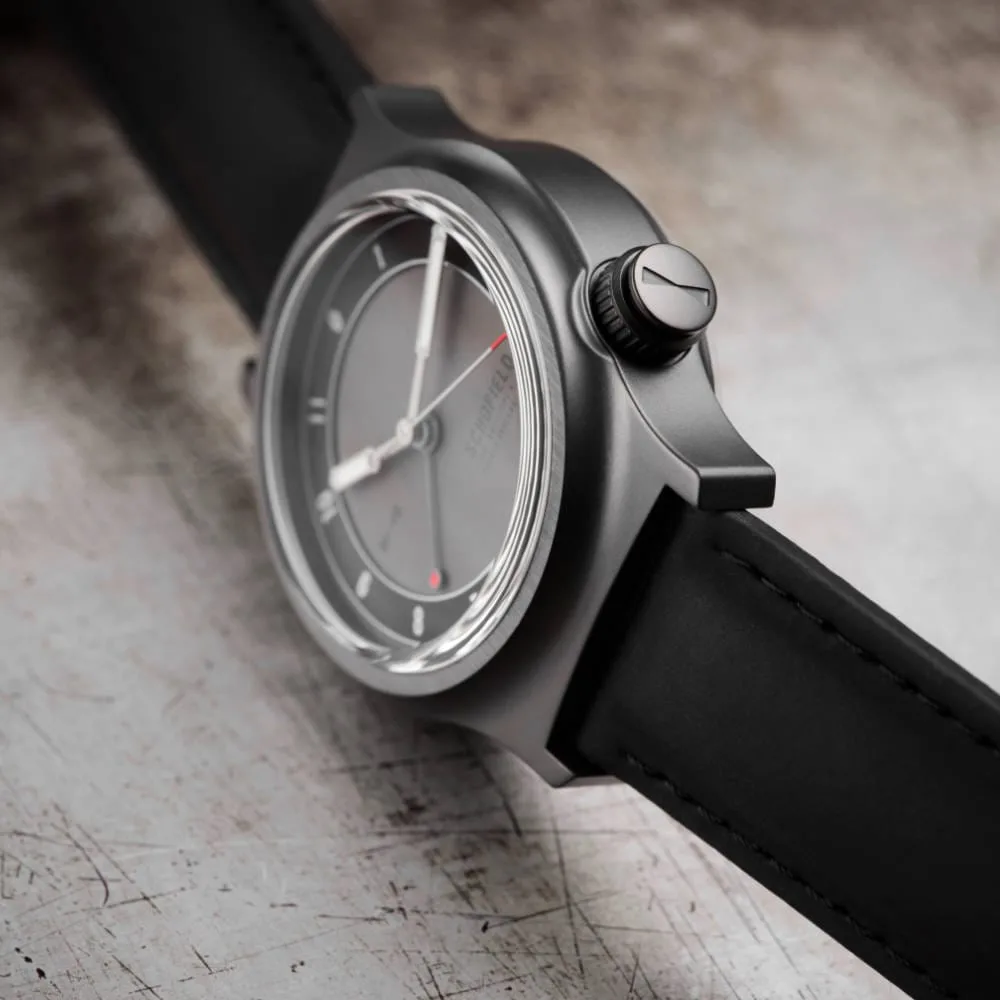 Close-up of a sleek, modern field watch known as Light ✦ The Dark One, featuring a dark metal case and a black leather strap. The gray watch face has minimalist design elements and subtle markings, with the crown adding a touch of elegance. The background is a textured, worn metal surface, enhancing the watch's contemporary aesthetic.