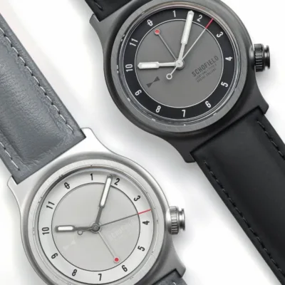 Two Schofield Light Field Watches displayed side by side on a white background. The top watch features a dark grey face with black and red accents, a black case, and a black leather strap. The bottom watch has a light grey face with dark grey and red accents, a light grey case, and a grey leather strap.