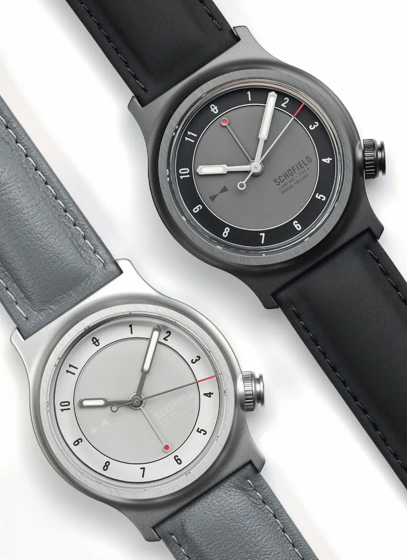 Two Schofield Light Field Watches displayed side by side on a white background. The top watch features a dark grey face with black and red accents, a black case, and a black leather strap. The bottom watch has a light grey face with dark grey and red accents, a light grey case, and a grey leather strap.