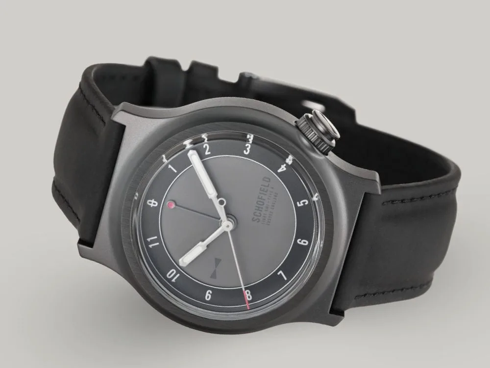 The Light - The Dark One watch is a sleek wristwatch featuring a matte grey dial with minimalist design elements. It has white luminescent hands, with a small red-tipped seconds and GMT hand. The watch has a black rubber strap and a DLC case with "SCHOFIELD" subtly printed on the dial.