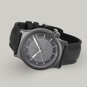 The Light - The Dark One watch is a sleek wristwatch featuring a matte grey dial with minimalist design elements. It has white luminescent hands, with a small red-tipped seconds and GMT hand. The watch has a black rubber strap and a DLC case with "SCHOFIELD" subtly printed on the dial.