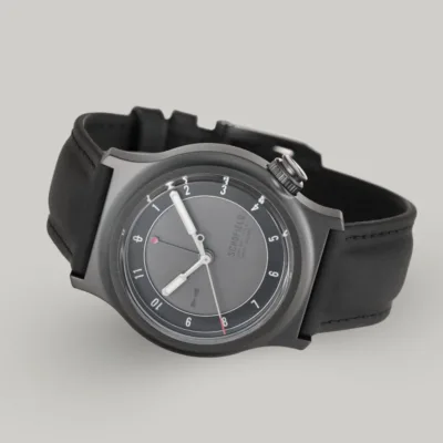 The Light - The Dark One watch is a sleek wristwatch featuring a matte grey dial with minimalist design elements. It has white luminescent hands, with a small red-tipped seconds and GMT hand. The watch has a black rubber strap and a DLC case with "SCHOFIELD" subtly printed on the dial.