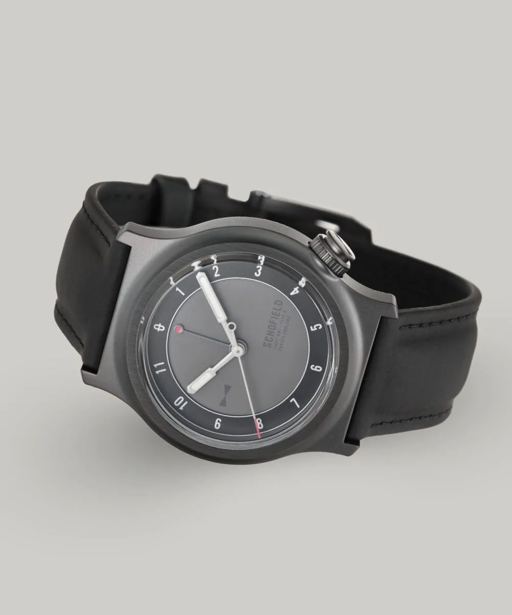 The Light - The Dark One watch is a sleek wristwatch featuring a matte grey dial with minimalist design elements. It has white luminescent hands, with a small red-tipped seconds and GMT hand. The watch has a black rubber strap and a DLC case with "SCHOFIELD" subtly printed on the dial.