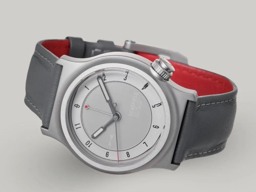 The Light One watch featuring a pale grey dial with minimalist design elements. A military watch. It has white luminescent hands, with a small red-tipped seconds and GMT hand. The watch has a grey leather strap and a bead blasted case with "SCHOFIELD" subtly printed on the dial.