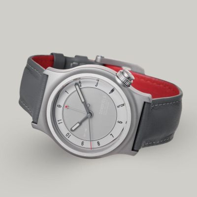 2025 PRODUCT TEMPLATE The Light One watch featuring a pale grey dial with minimalist design elements. A military watch. It has white luminescent hands, with a small red-tipped seconds and GMT hand. The watch has a grey leather strap and a bead blasted case with "SCHOFIELD" subtly printed on the dial.