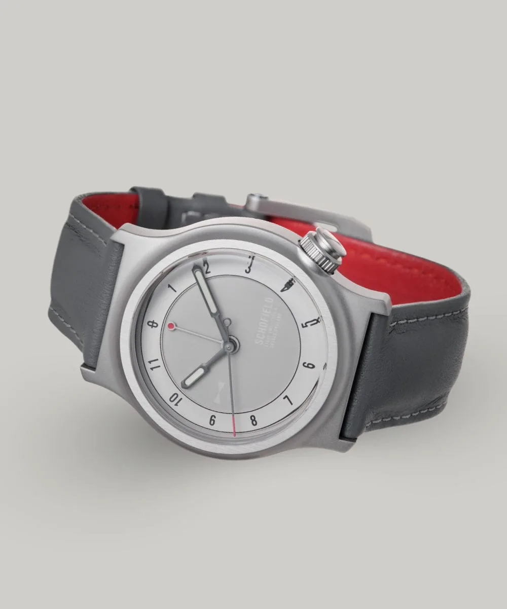 The Light One watch featuring a pale grey dial with minimalist design elements. A military watch. It has white luminescent hands, with a small red-tipped seconds and GMT hand. The watch has a grey leather strap and a bead blasted case with "SCHOFIELD" subtly printed on the dial.