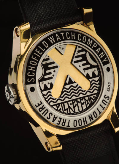 A close-up view of the back of a gold wristwatch from the Schofield Watch Company. The back features decorative engravings, including geometric patterns and the company's name. The watch also has a black leather strap and an intricate crown design.