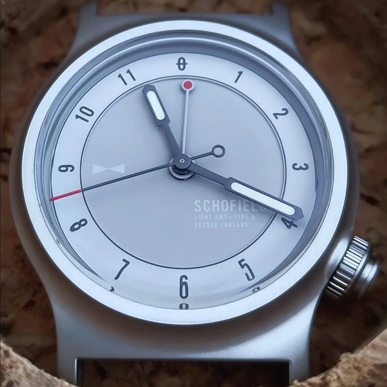 A close-up of a Schofield Light watch with a grey face and brushed bezel. The Light Field Watch, boasts minimalist design elements, including silver hour and minute hands and a red-tipped second and GMT hand. It is engraved with "Schofield Light GMT-Type A. Sussex England.