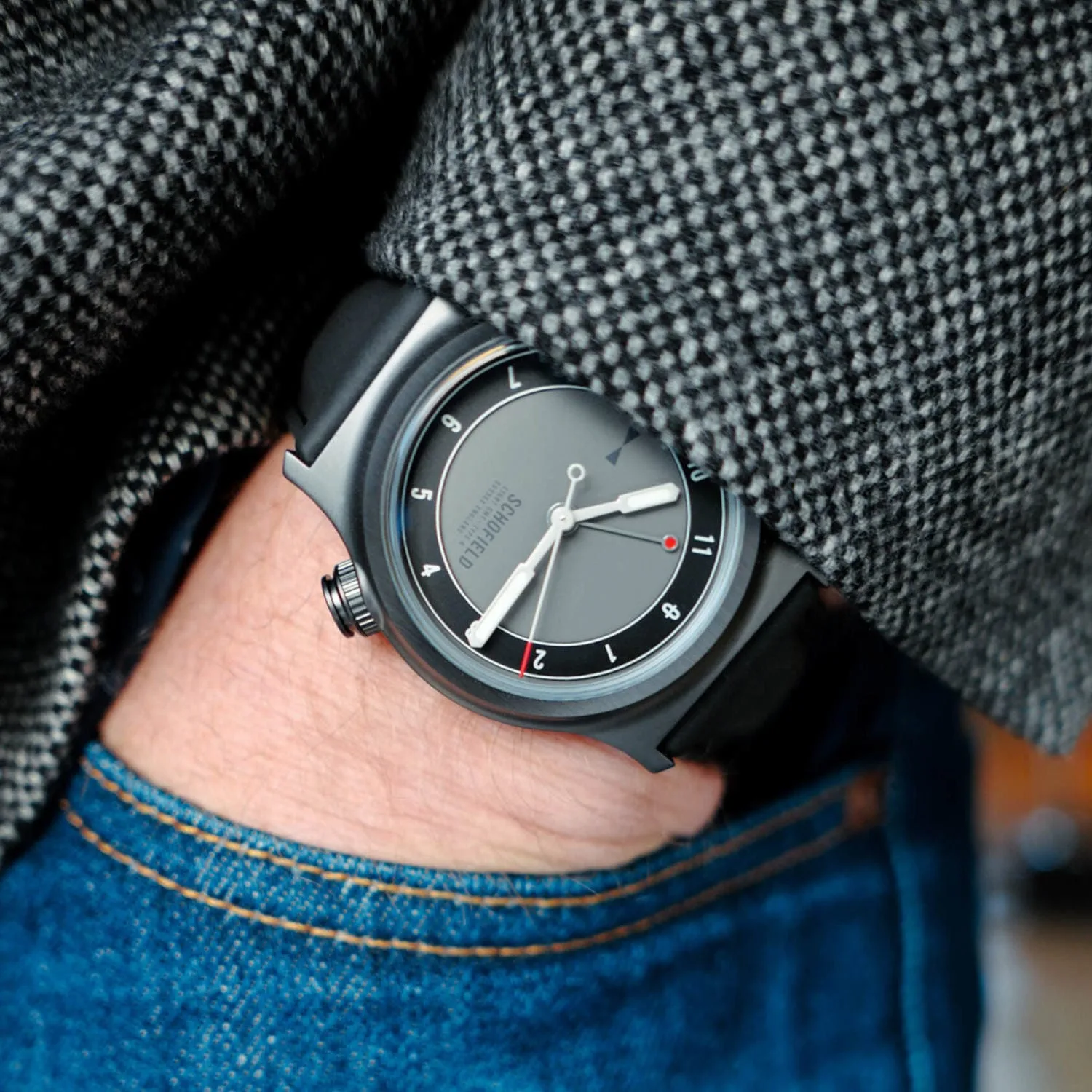 A person wearing blue jeans and a gray houndstooth-patterned jacket has their left hand in their pocket, revealing a stylish Schofield field watch with a black strap on their wrist.