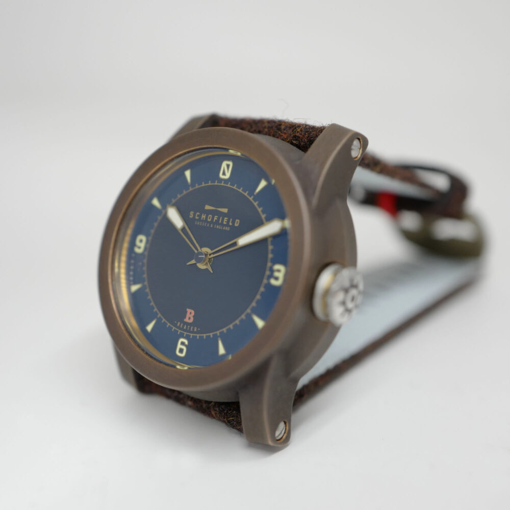 A Schofield Bronze watch featuring a navy blue face with brass details and a brown leather strap. The brand name "Schofield" and the lighthouse logo is prominently visible.