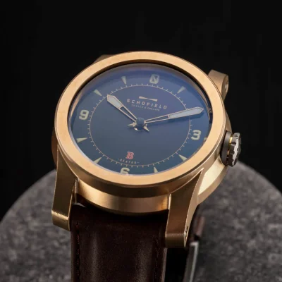 A raw bronze watch with a blue face, brown leather strap, and the brand name "Schofield" displayed on the dial.