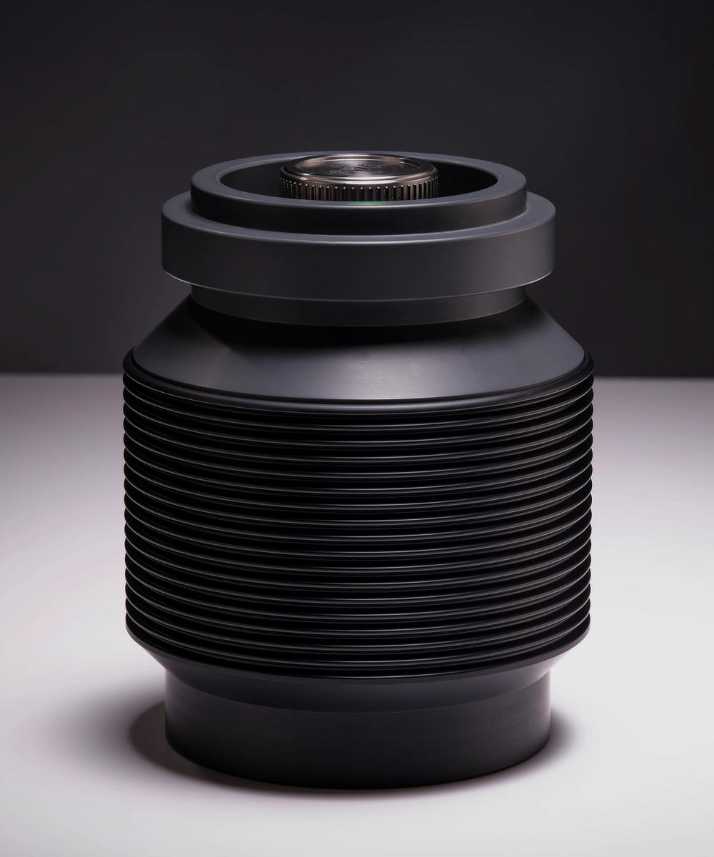 A modern, cylindrical black canister features multiple horizontal ridges and a smooth, flat top with a circular metallic component at the center. The dark background enhances the canister's sleek, industrial design.