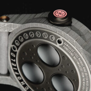 Close-up image of a Schofield Obscura with a textured grey strap and black and red crown. The case back features three circular sapphire crystals, a detailed case with various random markings, and an engraving that reads "Damascus steel, manual wind, pioneering watchmakers from the new era".