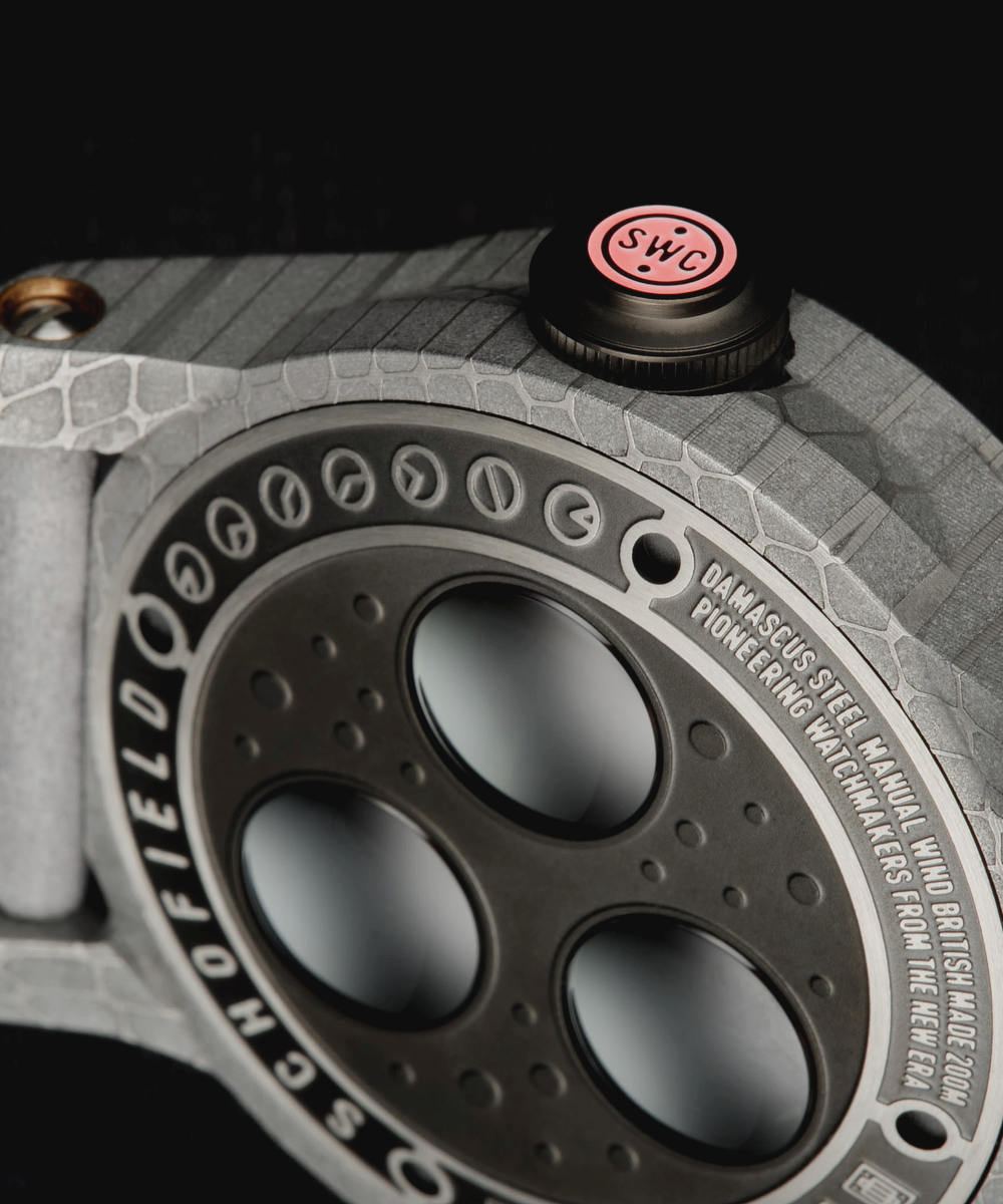 Close-up image of a Schofield Obscura with a textured grey strap and black and red crown. The case back features three circular sapphire crystals, a detailed case with various random markings, and an engraving that reads "Damascus steel, manual wind, pioneering watchmakers from the new era".