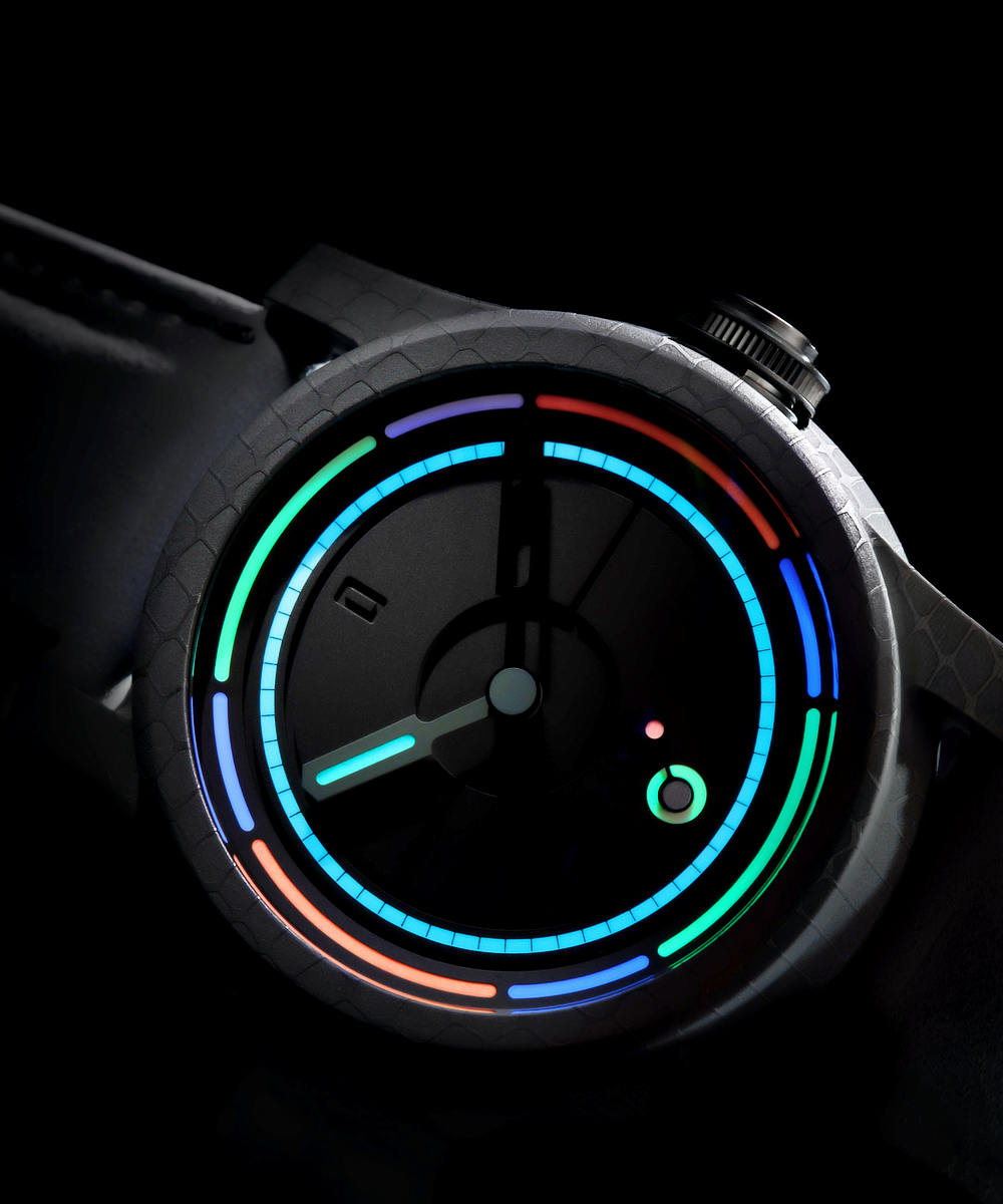 A close-up shot of a wristwatch with a dark face and a sleek, modern design. The watch features vibrant, colorful neon-like light accents in blue, green, red, and orange around the perimeter and in circular patterns on the watch face against a black background.