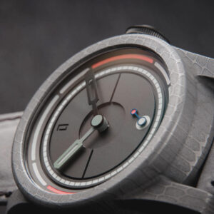 Close-up image of a modern wristwatch with a minimalist design. The watch has an Obscure matte black dial with green hour and black minute hands, a small red and blue second hand. The textured grey case and strap enhance its sleek, contemporary look.