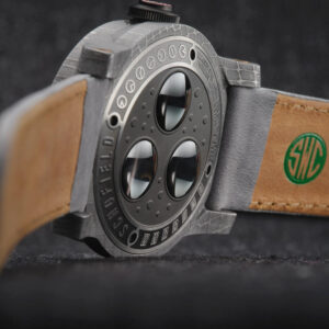 The underside of a wristwatch with a grey textured obscura case back and a leather strap. The watch features three circular windows on the back and the strap has a green logo with the letters "SWC" embossed.