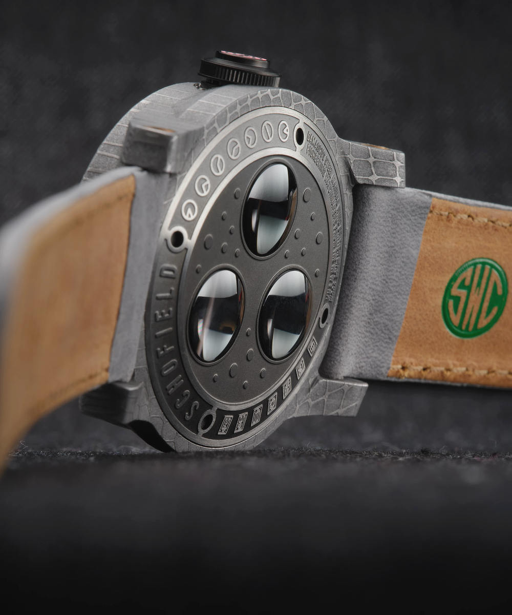 The underside of a wristwatch with a grey textured obscura case back and a leather strap. The watch features three circular windows on the back and the strap has a green logo with the letters "SWC" embossed.