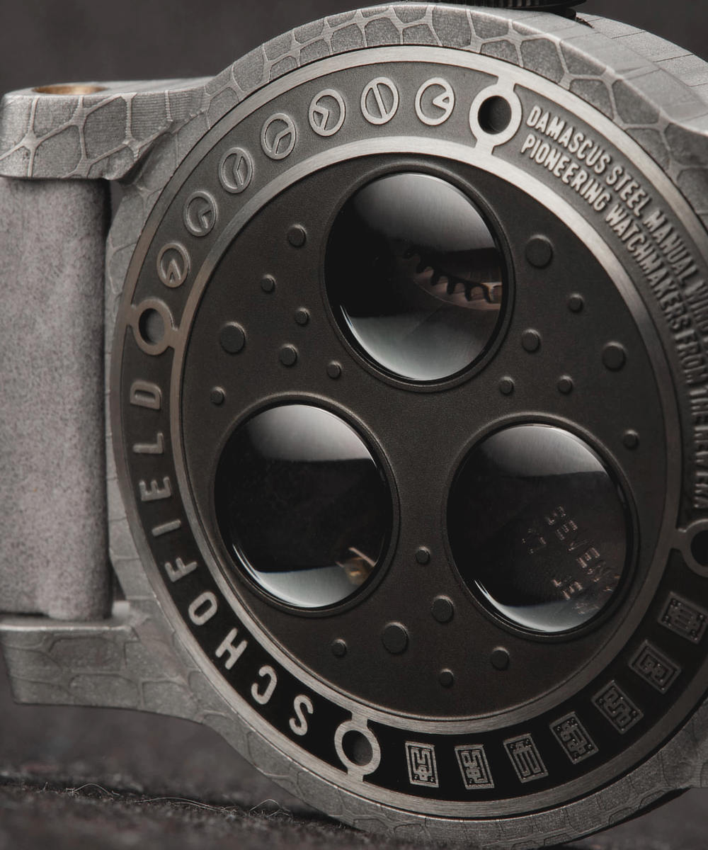 Close-up of a luxury wristwatch showcasing a round, textured, matte grey case with three large circular windows on the back. Engraved text on the caseback rim includes "DAMASCUS STEEL" and "PIONEERING WATCHMAKING".