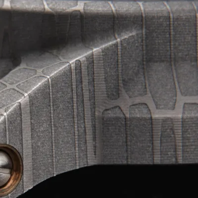 Close-up of an intricately etched metallic surface with a textured, grid-like pattern. The British-made watch case has a matte finish and features a bronze bushed screw partially visible in the lower left corner.