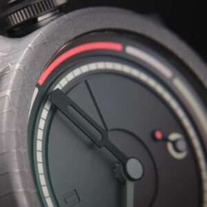 Close-up of a modern Obscura watch by Schofield. The face is predominantly black with red, white, and grey accents. The design is minimalistic with a small seconds sub-dial, and textured casing.