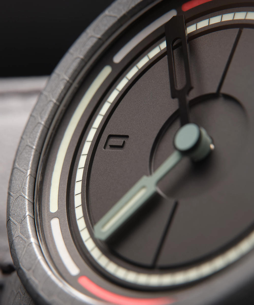 Close-up of a British-made modern analog wristwatch with a dark face. The watch features thick, stylized hands and luminous hour markers.