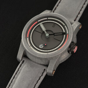 A close-up of a stylish Schofield wristwatch with a grey textured strap resting on a dark surface. The Obscura watch face features a minimalistic design with black and white hour, minute, and second hands, along with a red marker near the edge and some intricate inner circle details.