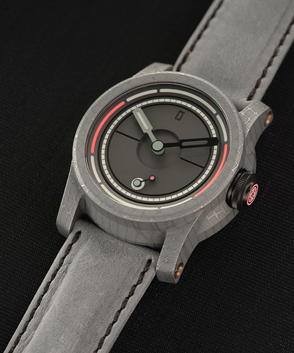 A close-up of a stylish Schofield wristwatch with a grey textured strap resting on a dark surface. The Obscura watch face features a minimalistic design with black and white hour, minute, and second hands, along with a red marker near the edge and some intricate inner circle details.