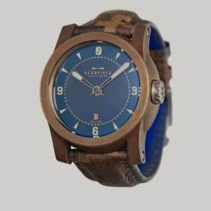 The B3 ✦ Patinated Bronze wristwatch with a blue dial and brown leather strap. The watch face features bold luminescent numerals at 3, 6, 9, and 12, with smaller indicators for other hours and minutes. The brand name "Schofield" is visible on the dial, along with the Beam of light logo.