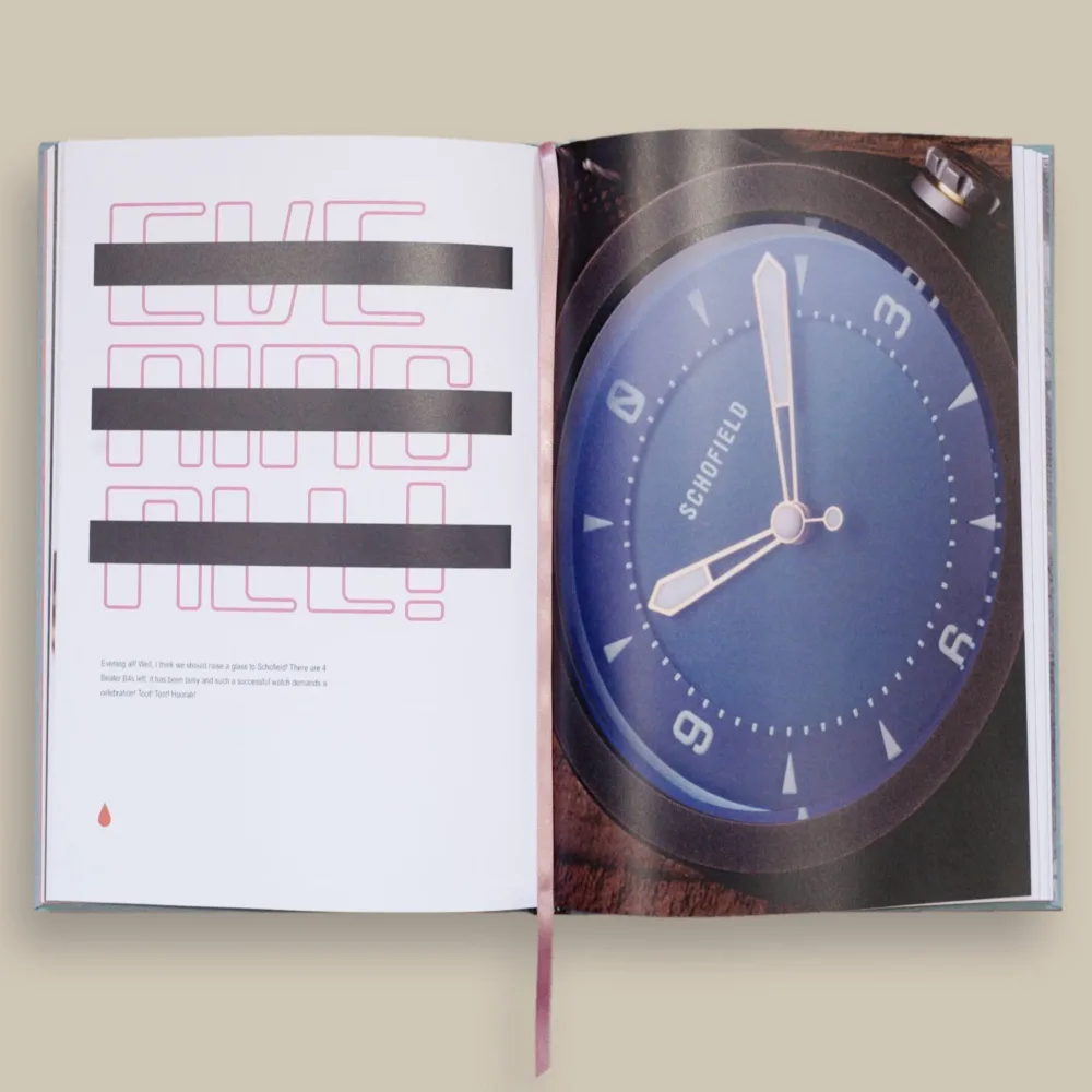 Open Six Pips book showing graphic text and a blue Schofield watch face. A pink ribbon bookmark is draped over the pages.