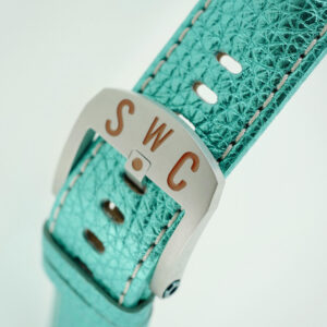 Close-up of a turquoise metalic watch strap with a Schofield buckle featuring the initials "SWC" engraved in red. The silver buckle complements the textured design of the strap, and several adjustment holes are visible.