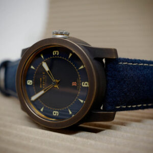 Close-up of a person holding a bronze watch case between their thumb and index finger. The focus is on the steel crown and the cylindrical shape of the case, made of Bronze.