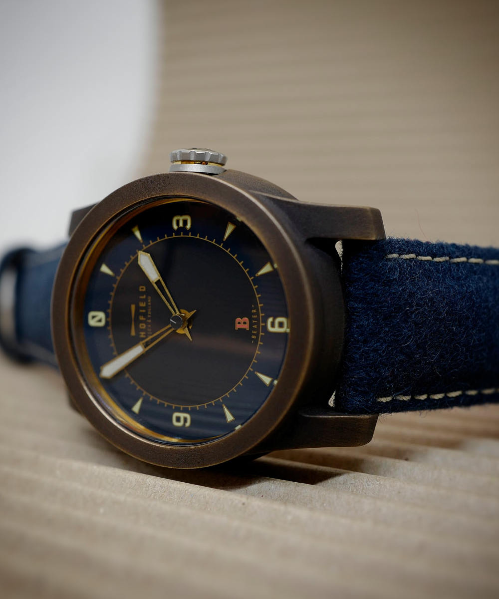 Close-up of a person holding a bronze watch case between their thumb and index finger. The focus is on the steel crown and the cylindrical shape of the case, made of Bronze.