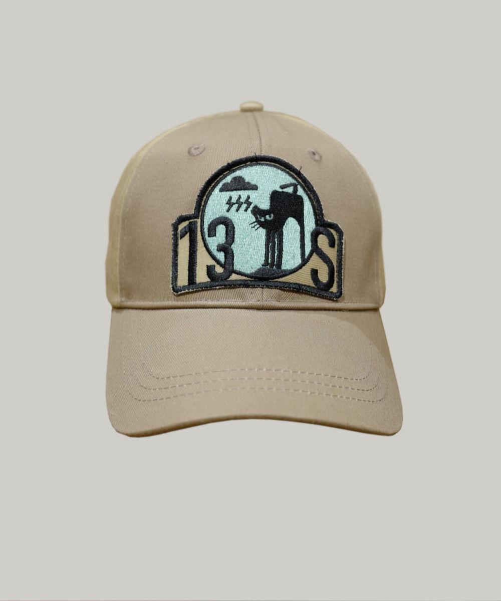 A BAD MOOD CAP in beige showcases a circular patch adorned with a stylized figure, the number 130S, and abstract black and teal designs. The cap is set against a simple light gray background, enhancing its minimalist appeal.