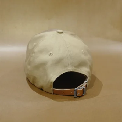 2024 PRODUCT TEMPLATE On a matching beige surface with a neutral background, the beige BAD MOOD CAP is displayed from the back. It features an adjustable strap with a metal buckle, highlighting its understated style.