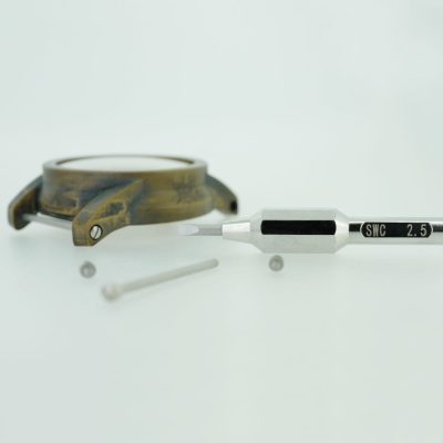 Next to a watch case with strap bars, a screwdriver labeled "SWC 2.5" lies on a white surface, alongside a separate metal screw.