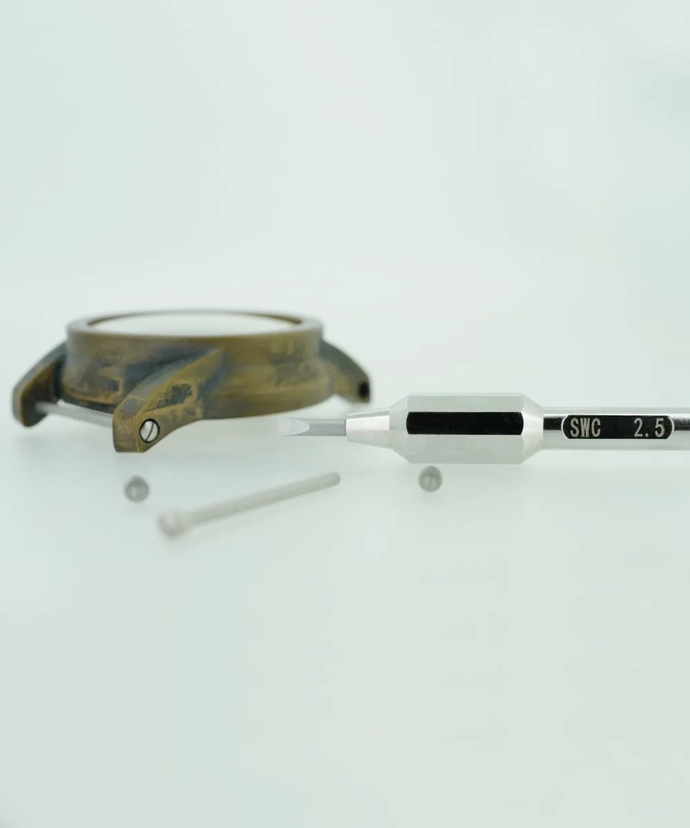 Next to a watch case with strap bars, a screwdriver labeled "SWC 2.5" lies on a white surface, alongside a separate metal screw.