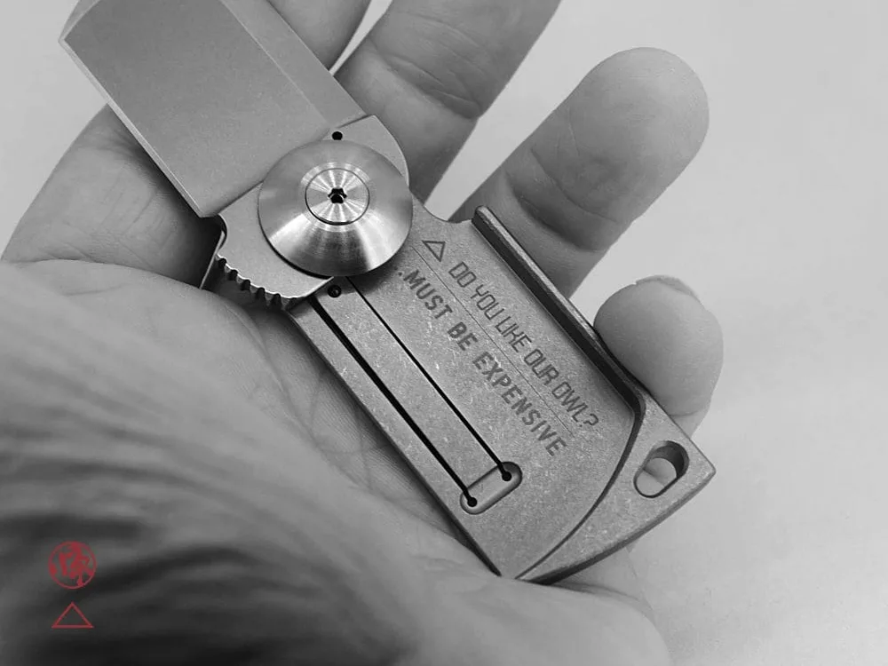A hand holds a compact, rectangular folding knife labeled “MUST BE EXPENSIVE.” The knife has an industrial design and engraved details. The image is in black and white with graphic elements and text at the bottom.