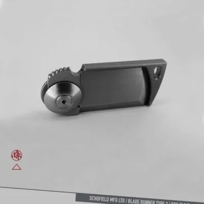 A compact, rectangular metallic object with a circular pivot and ridged edge, photographed on a light background. Branding and text at bottom right read: "SCHOFIELD MFG LTD / BLADE RUNNER TYPE 2 / DOG TAG Ti.