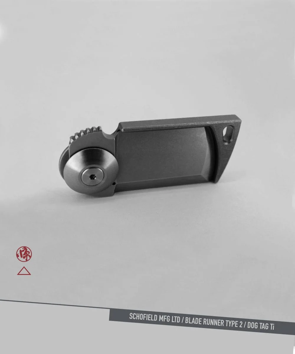 A compact, rectangular metallic object with a circular pivot and ridged edge, photographed on a light background. Branding and text at bottom right read: "SCHOFIELD MFG LTD / BLADE RUNNER TYPE 2 / DOG TAG Ti.