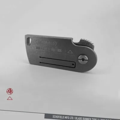 A compact, metal folding knife resembling a dog tag, engraved with "SCHOFIELD" and various text codes, displayed on a plain gray background with subtle branding and product name at the bottom.