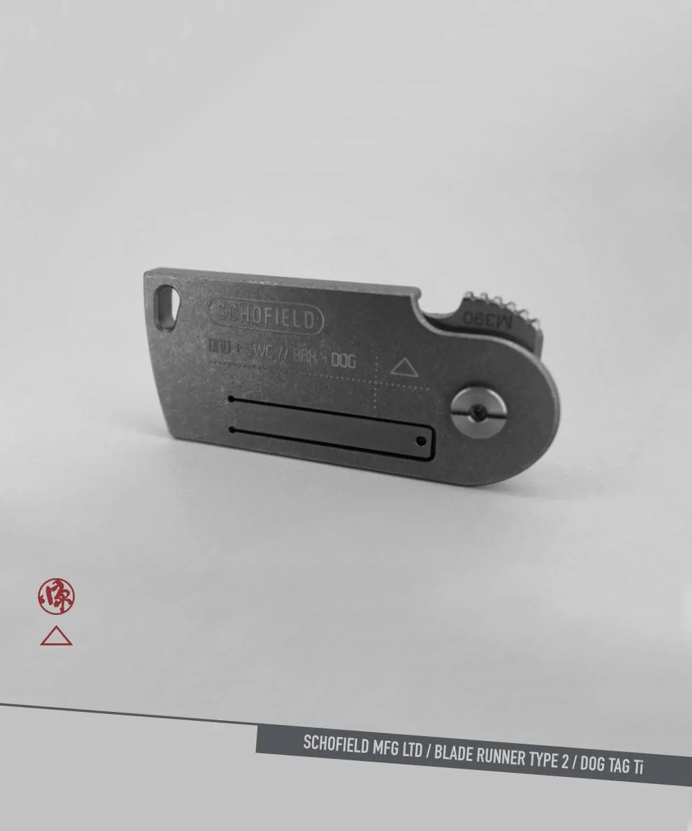 A compact, metal folding knife resembling a dog tag, engraved with "SCHOFIELD" and various text codes, displayed on a plain gray background with subtle branding and product name at the bottom.
