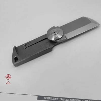 A compact, gray folding knife with a sleek, minimalist design is displayed on a light gray surface. Text reads "SCHOFIELD MFG LTD / BLADE RUNNER TYPE 2 / DOG TAG Ti" in the bottom right corner.